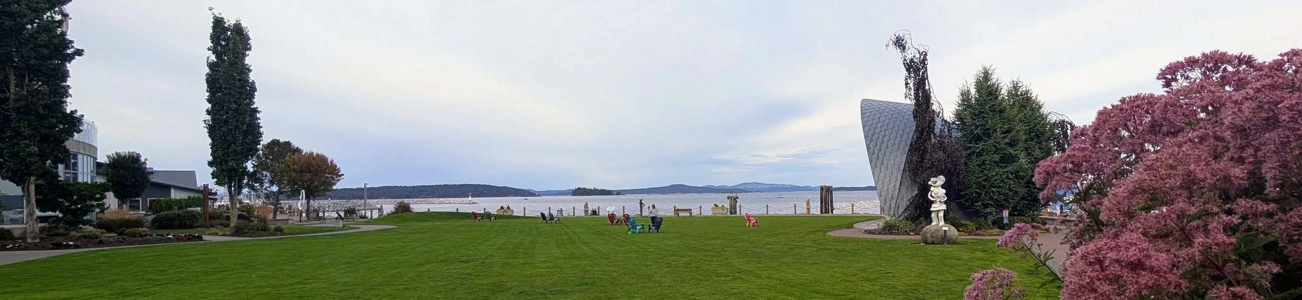 A waterfront park lawn in Sidney, with public art, a flowering shrub, and the water beyond