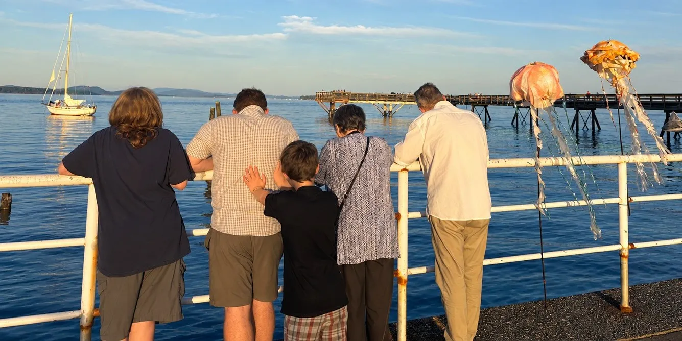Erin's family, seen from behind, leaning on a waterfront railing at sunset watching a sailboat, with the Sidney pier in the background